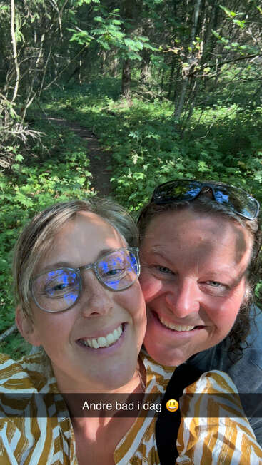 Duas pessoas sorridentes tiram uma selfie alegre em uma floresta verdejante. A luz do sol passa por entre as árvores. A legenda diz: "Andre bad i dag 😀".