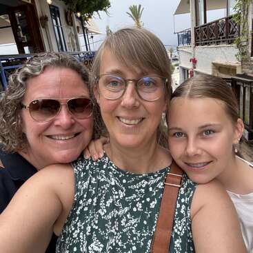 Tres personas sonrientes, probablemente una familia, posan muy juntas al aire libre. Dos adultos llevan gafas, uno con gafas de sol. La joven lleva ortodoncia. Parecen felices y relajados.