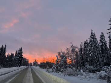 Une route enneigée s'étend dans une forêt hivernale, avec des arbres recouverts de neige de chaque côté. Le ciel est illuminé par un coucher de soleil orange et violet éclatant.