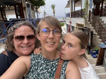 Three people—two adults and a child—smile and pose for a selfie on a sunny street with Mediterranean buildings, outdoor stairs, and tropical palm tree views.