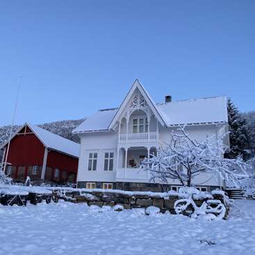 Una encantadora casa blanca con intrincados adornos se alza junto a un granero rojo. Ambos edificios están rodeados de nieve y árboles bajo un claro cielo azul de invierno.