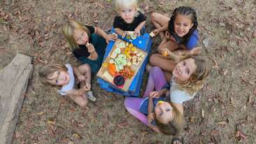 Six children sit on the ground outdoors, gathered around a small table with assorted fruits and snacks, smiling up at the camera, surrounded by dry leaves.
