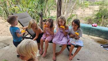 Six children sit outdoors, some on a lounge chair, enjoying slices of fruit. One child reads a book. Greenery and sandy landscape surround the relaxed, joyful scene.