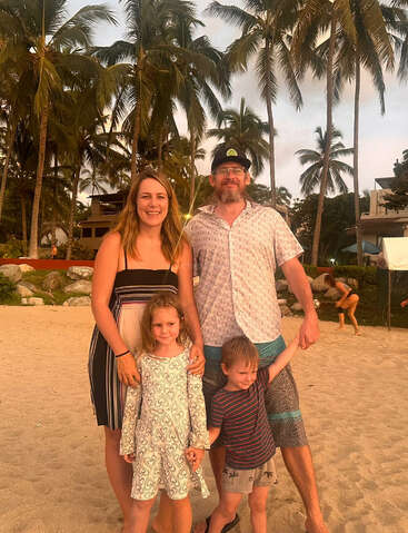 A happy family of four poses on a sandy beach at sunset, surrounded by tall palm trees, enjoying a tropical vacation with warm, golden lighting.