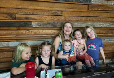 A woman sits with five young children, each displaying different expressions. They are indoors, in front of a wooden wall, sharing a casual, candid moment together.