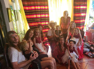 A family group poses indoors with colorful curtains and gifts. Most wear red or white dresses, and some children sit on adults’ laps, smiling happily together.