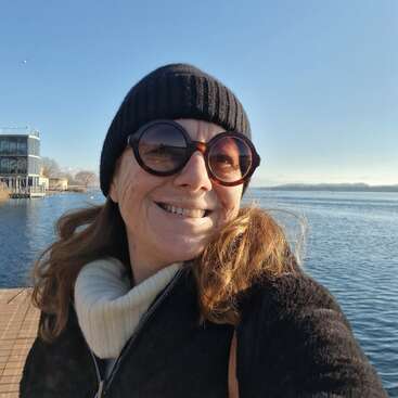L'image représente une femme debout sur un quai, face à l'appareil photo, avec un plan d'eau serein et un ciel bleu clair en arrière-plan. Elle est chaudement vêtue.