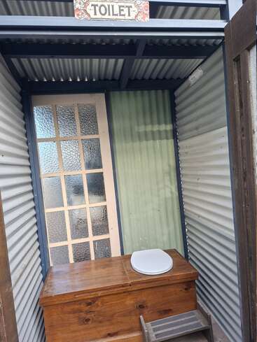 Cette photo montre des toilettes extérieures à l'ancienne, avec un siège en bois, des murs et un toit en tôle ondulée, un tabouret et une pancarte « TOILET » au-dessus de l'entrée.