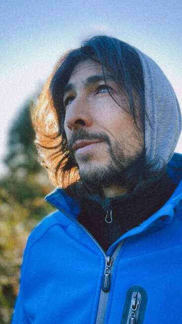 A man with long hair and a beard, wearing a blue jacket and gray hood, gazes thoughtfully into the distance, bathed in soft outdoor sunlight.
