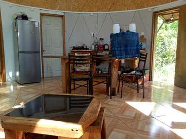 This image shows a bright rustic kitchen with a wooden table, chairs, refrigerator, and various kitchen appliances near an open door leading outside to greenery.