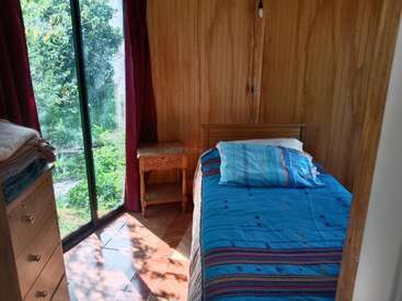 A small, cozy bedroom with wooden walls, a single bed with blue bedding, a bedside table, dresser, and large window overlooking a green garden.