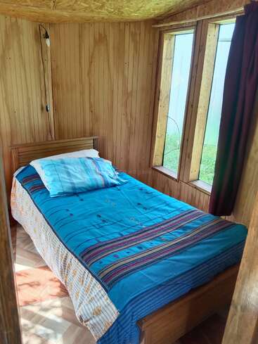 A small, cozy wooden room features a single bed with a bright blue blanket, patterned bedspread, and pillow. Sunlight streams through three tall windows.