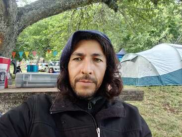 Ein Mann mit langen Haaren und einem Bart, der eine schwarze Jacke und eine Kapuze trägt, macht ein Selfie im Freien. Zelte, bunte Fahnen und Bäume schmücken den Zeltplatz.