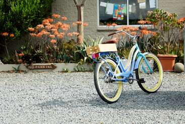 A imagem mostra uma bicicleta estacionada em frente a uma casa, com um gramado e árvores visíveis ao fundo. A bicicleta está posicionada em uma entrada de automóveis ou estrada pavimentada.