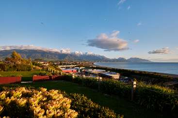 Uma bela cidade costeira com casas coloridas, jardins exuberantes e flores desabrochando, com vista para um mar sereno e montanhas cobertas de neve sob um céu azul claro.