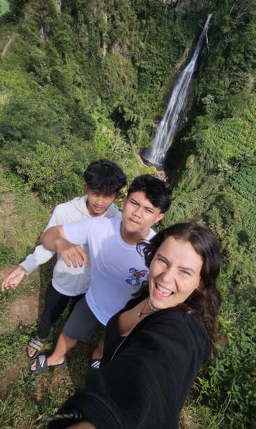 Three friends take a cheerful selfie on a lush green cliff, with a beautiful tall waterfall cascading down amidst dense forest in the background.