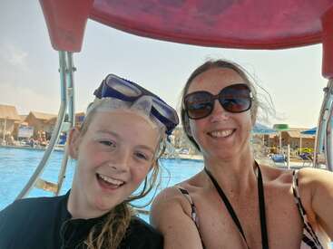 Eine Frau und ein Mädchen lächeln für ein Selfie an einem Pool. Das Mädchen trägt eine Schwimmbrille auf dem Kopf; beide wirken glücklich und entspannt und genießen den Sommer.