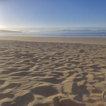 Ein ruhiger Sandstrand erstreckt sich bis zu den sanften Wellen und den fernen Bergen, badet im sanften Morgenlicht unter einem strahlend blauen Himmel und schafft eine friedliche Atmosphäre.