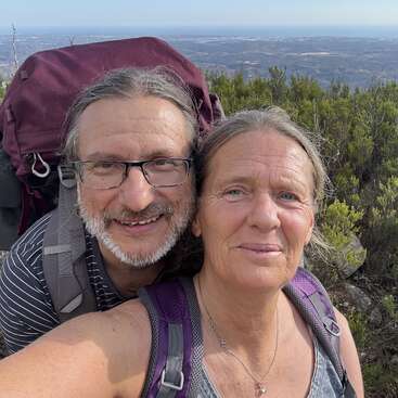 A smiling couple, both wearing backpacks, take a selfie during a hike. Behind them, lush green bushes and a distant scenic landscape are visible.