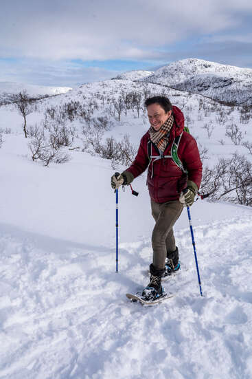 A person, dressed warmly in a red jacket and scarf, hikes through snowy mountains with snowshoes and trekking poles, surrounded by leafless trees under a cloudy sky.