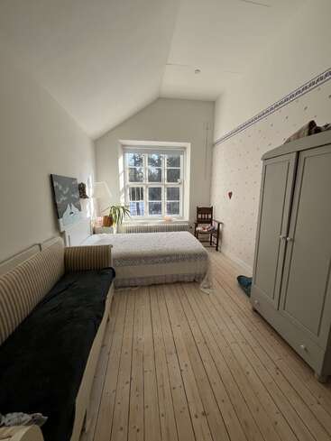 A cozy bedroom with wooden floors, bed near a large window, striped sofa, wardrobe, chair, plant, lamp, wall art, and patterned wallpaper. Daylight fills the room.