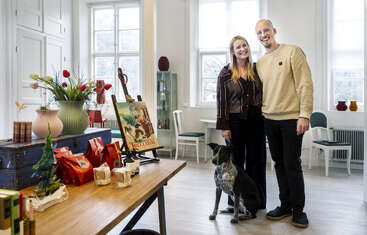 A smiling couple stands together in a bright, cozy room with large windows. A black-and-white dog sits at their feet, and art is displayed nearby.