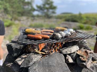 A rustic outdoor barbecue setup with sausages, corn, and foil-wrapped food grilling over an open fire on a stone grill, surrounded by nature.