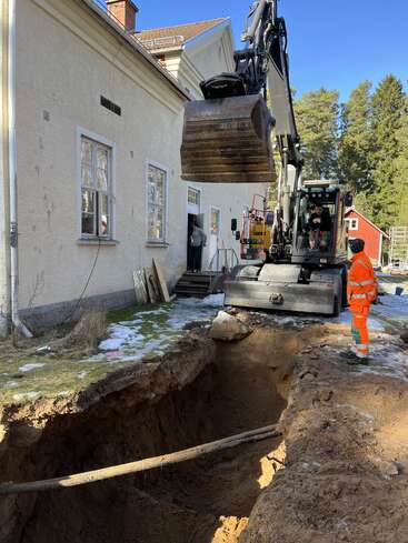 An excavator is digging a large trench next to a house. A worker in orange safety gear observes while another stands near the building entrance.