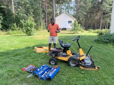 A man stands on green grass near a yellow riding lawnmower, various tools spread out. Surrounded by trees and a small white house in the background.