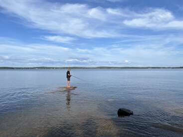 A person stands on a shallow sandbar in a calm lake, fishing under a vast blue sky with scattered clouds, enjoying a peaceful, serene moment.