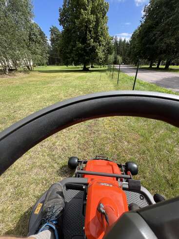 The image shows a person operating a red lawnmower on a sunny day, cutting grass in a spacious yard surrounded by tall green trees and blue sky.