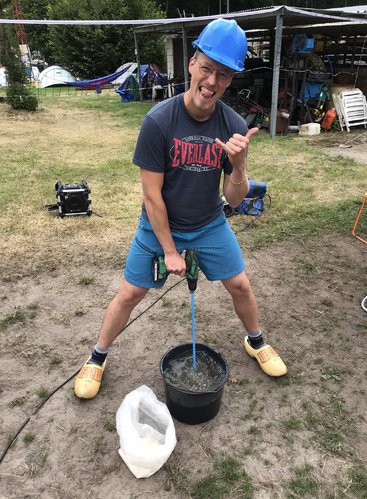 Ein Mann mit blauem Schutzhelm und Holzschuhen mischt draußen mit einer Bohrmaschine Zement in einem Eimer, lächelt dabei und streckt den Daumen hoch.