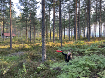 Eine Frau lächelt und winkt mit einem roten Gegenstand in einem üppigen, grünen Wald. Hohe Kiefern umgeben sie, im Hintergrund ist eine Holzhütte zu sehen.