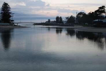 Un río tranquilo refleja el cielo nublado del atardecer, con siluetas de árboles y casas a ambos lados, y un puente que se extiende a lo lejos.