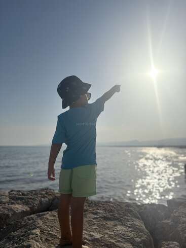 A child stands on rocky shore, wearing a hat and sunglasses, pointing towards the bright sun over the sparkling sea, embracing a sunny, adventurous day.