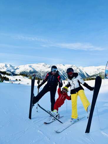 A family of three poses cheerfully on a snowy ski slope, holding hands. Snow-capped mountains and a clear blue sky create a stunning alpine backdrop.