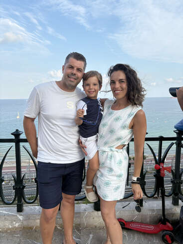 A smiling family of three poses by a seaside railing. The parents hold their child between them. A red scooter rests nearby. Beautiful blue sky above.