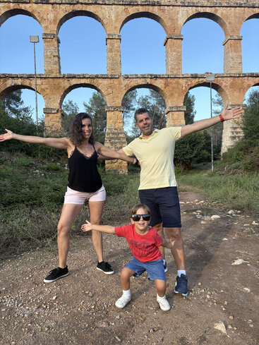 A happy family poses in front of an ancient stone aqueduct, arms stretched wide, enjoying a sunny day outdoors, with greenery and blue sky surrounding them.