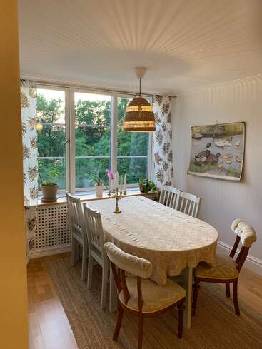 This cozy dining room features a lace-covered table, white chairs, patterned curtains, plant-filled windowsill, wicker lamp, nature painting, and leafy green view outside.