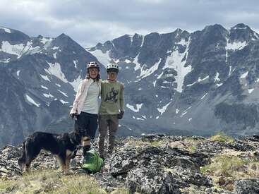 Two people wearing helmets and outdoor clothing stand on rocky terrain with a dog. Snow-capped mountains and a cloudy sky form the stunning, dramatic background.