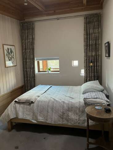 This cozy bedroom features a neatly made bed, wooden ceiling, small windows, simple artwork, bedside table with a clock, and soft natural light, creating a peaceful atmosphere.