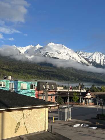 A small town sits quietly under clear blue skies, colorful buildings, and power lines. Snow-capped mountains loom majestically in the background with mist and greenery below.