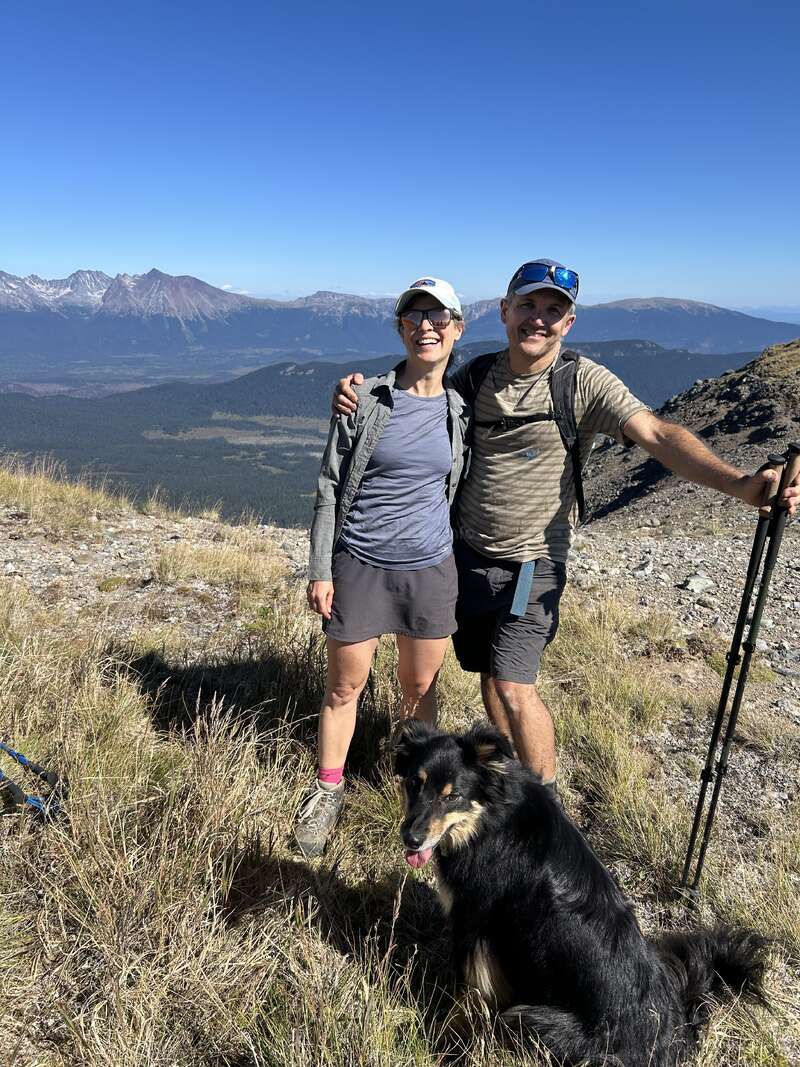 A smiling couple stands arm-in-arm on a mountain trail, hiking poles in hand. Their black dog sits happily at their feet amid breathtaking scenic views.
