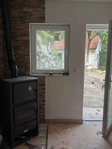 Ein gemütliches Interieur mit einem schwarzen Holzofen neben einem Fenster und einer offenen Tür, mit Blick auf eine Außenlandschaft mit Felsen und einem kleinen rustikalen Haus.