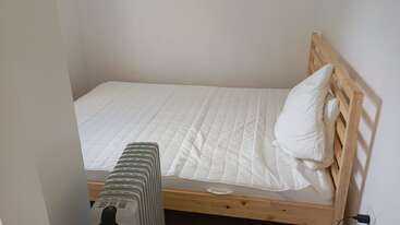 Das Bild zeigt ein kleines, einfaches Schlafzimmer mit einem Bett aus Holz, einer weißen Matratze, einem Kissen, weißer Bettwäsche und einem tragbaren Heizgerät daneben.