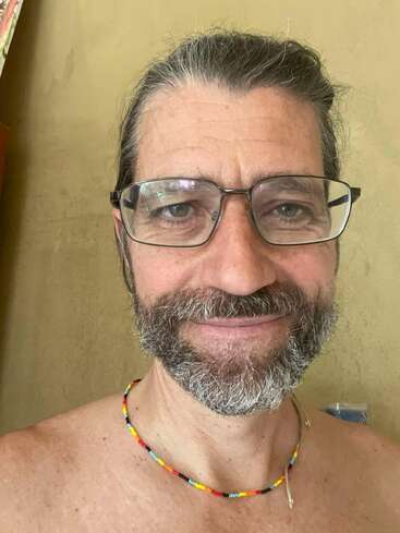Ein Mann mittleren Alters mit Brille und grauem Bart lächelt warmherzig. Er trägt eine bunte Perlenkette, ist ohne Hemd und steht an einer schlichten Wand.