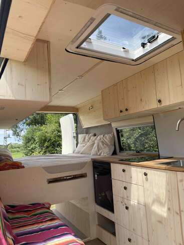Dieses Bild zeigt ein gemütliches, modernes Wohnmobil mit hellen Holzschränken, einem Dachfenster, einem Bett, einer kompakten Küche, farbenfrohen Sitzgelegenheiten und Panoramafenstern mit Blick auf üppiges Grün draußen.