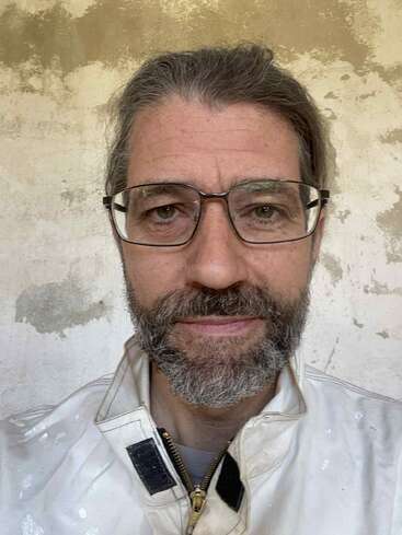Ein Mann mittleren Alters mit grauen Haaren, einer Brille und einem Bart trägt eine weiße Jacke. Er steht vor einer strukturierten, cremefarbenen Wand und schaut direkt in die Kamera.