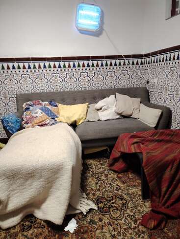 Ein gemütliches, unordentliches Wohnzimmer mit einem gemusterten Teppich, einem grauen Sofa, vielen Kissen und Decken. Dekorative Kacheln säumen die Wände, über denen ein helles blaues Licht hängt.