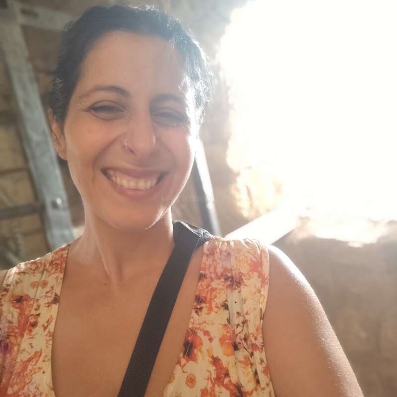 Eine lächelnde Frau macht ein Selfie in weichem, warmem Licht. Sie trägt ein geblümtes Kleid, hat einen Nasenring und steht vor einem hellen, sonnenbeschienenen Hintergrund.